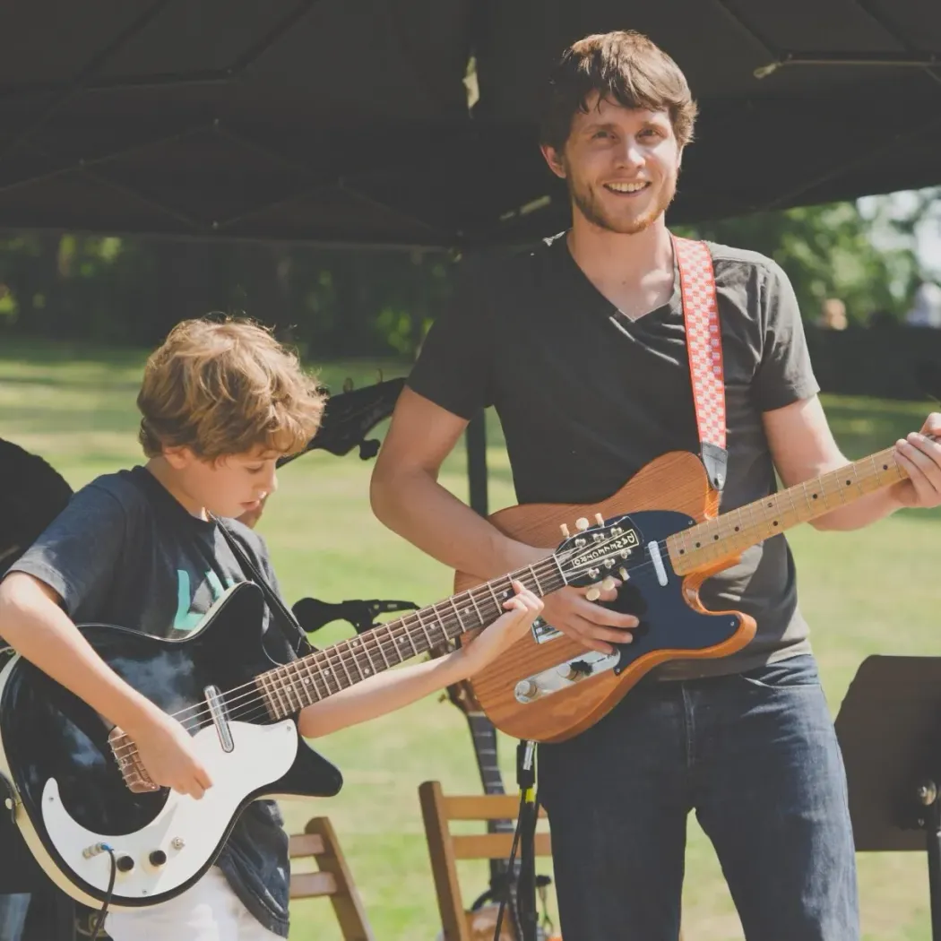 Founder Alex Stanton plays guitar on an outdoor stage alongside a young student