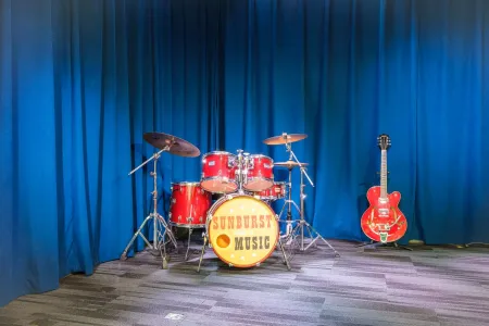 A red drum kit with the Sunburst School of Music logo and a hollow-body guitar on stage in front of blue curtains