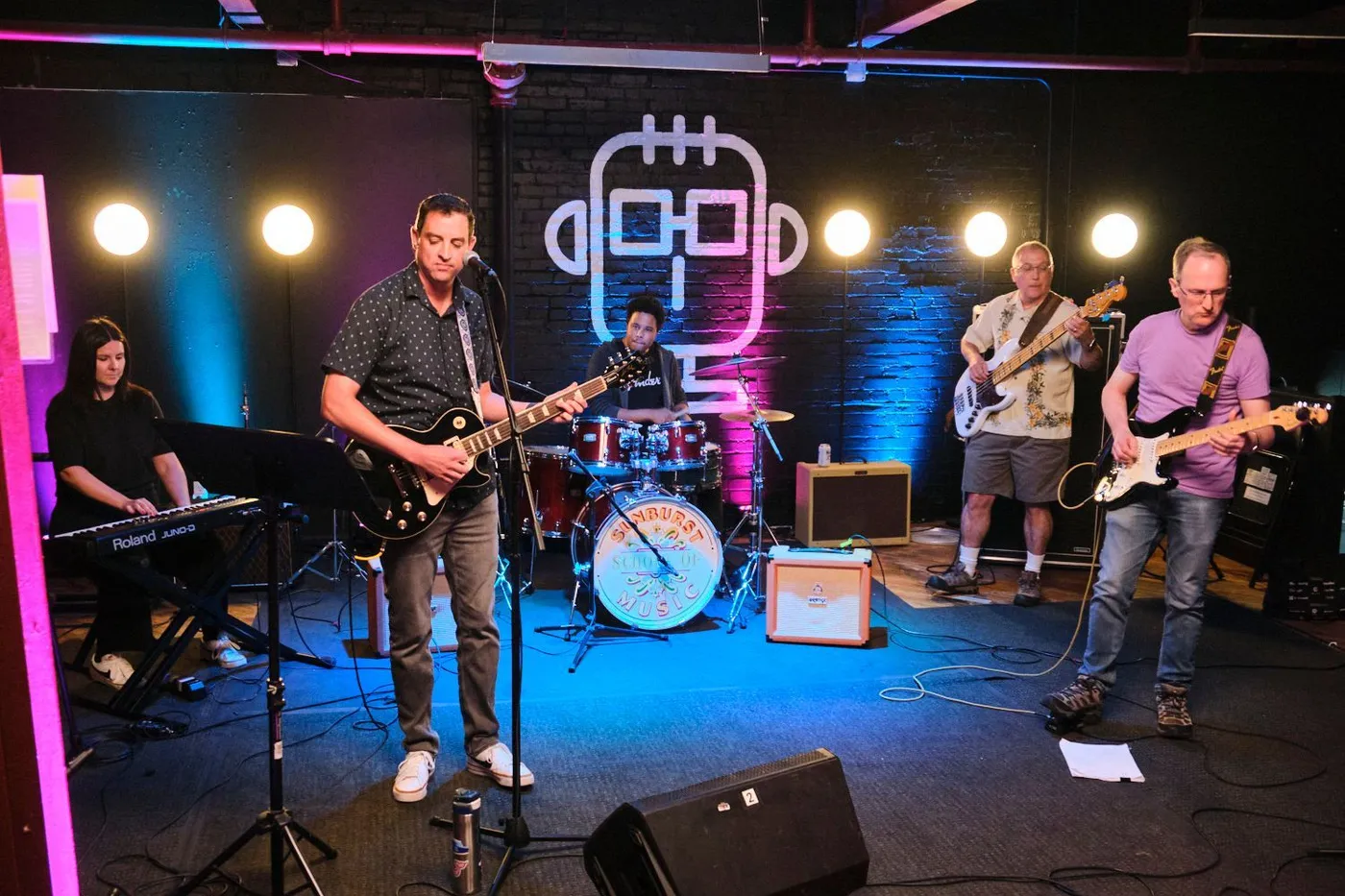 An adult band performs under stage lights in front of the Sunburst logo