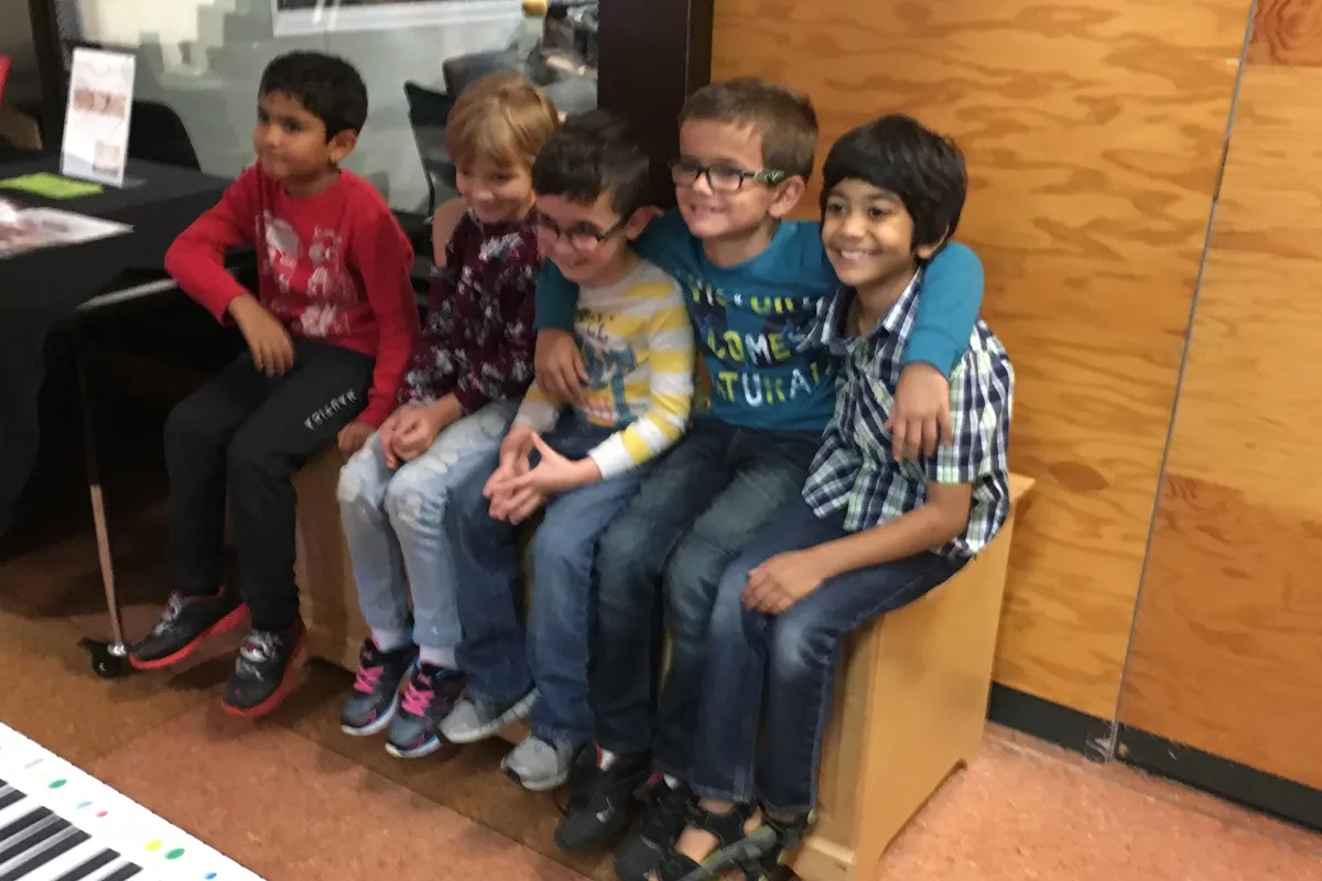 Five young students sit squeezed together on a bench beside a giant floor keyboard