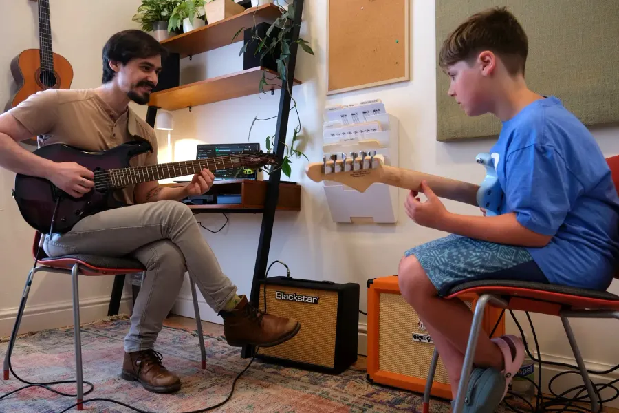 A teacher and a young student play electric guitars through amps in a lesson room