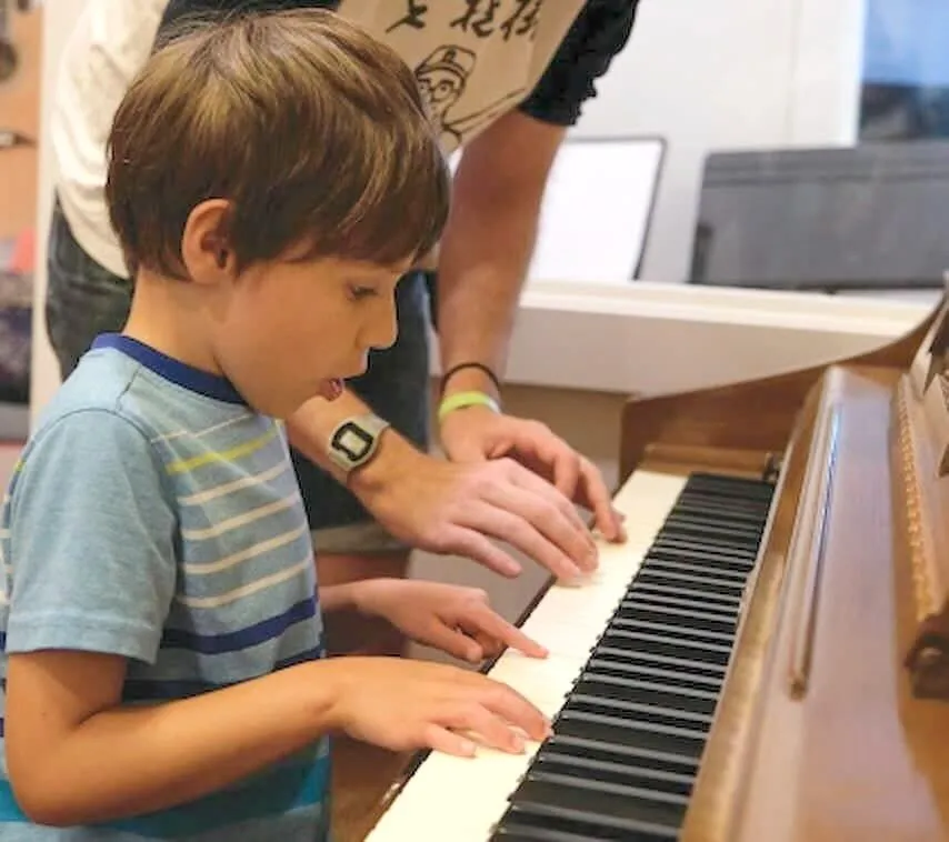 A young beginner picks out notes at an upright piano with his teacher standing by